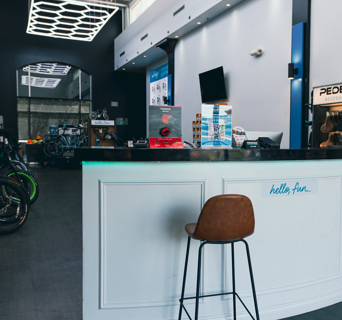 Pedego- A modern bike shop reception area with a white front desk, a brown chair, and cycling accessories displayed on the counter. The shop has black walls and a unique geometric ceiling light.