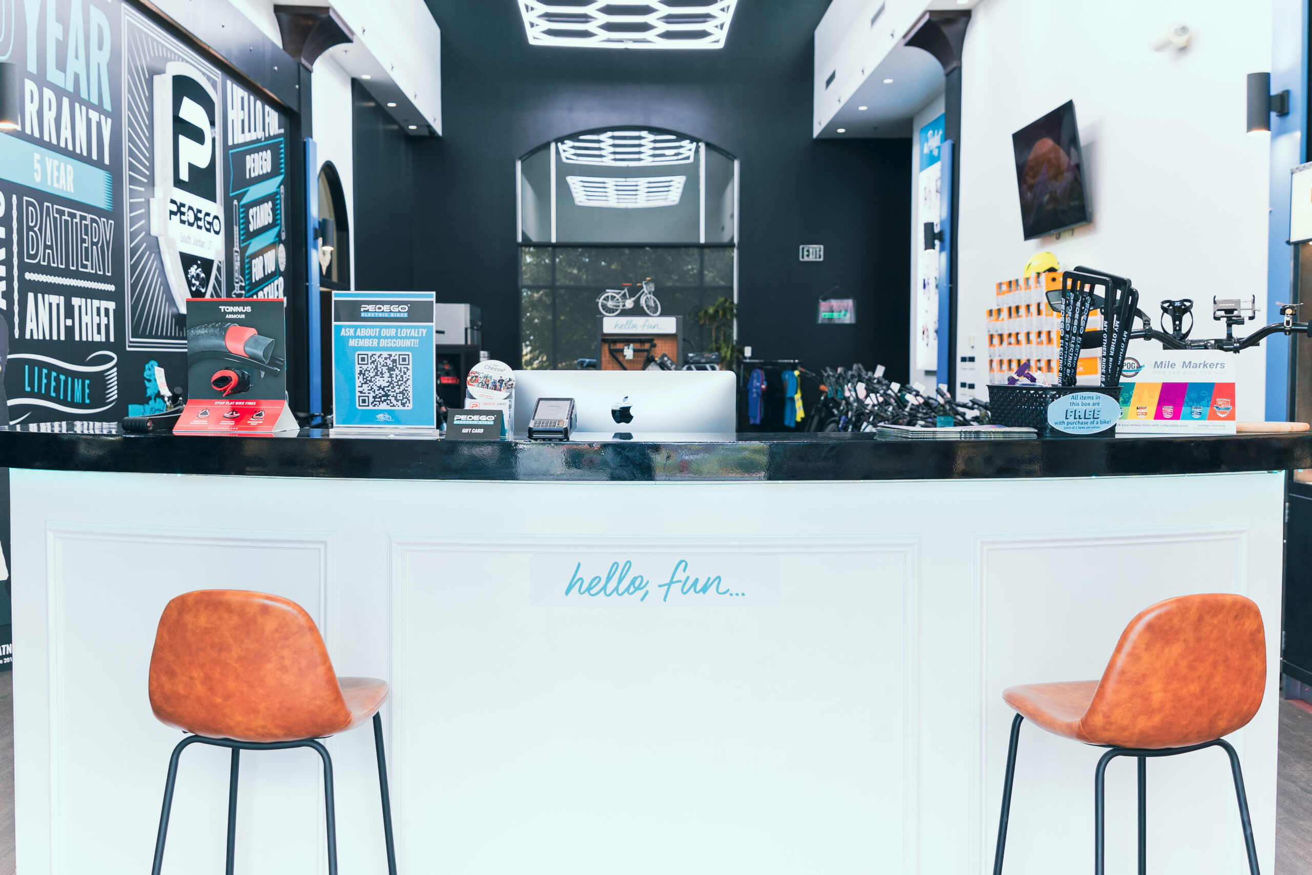 Pedego- A modern bike shop reception area with a counter, computer, two orange chairs, and various cycling accessories displayed. The wall has promotional posters and a bike hanging above.